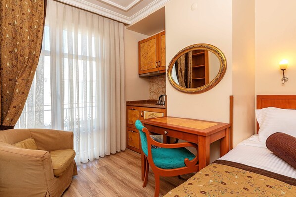 Habitación de hotel con cama, escritorio de madera, silla, zona de cocina y un ventanal con cortinas.