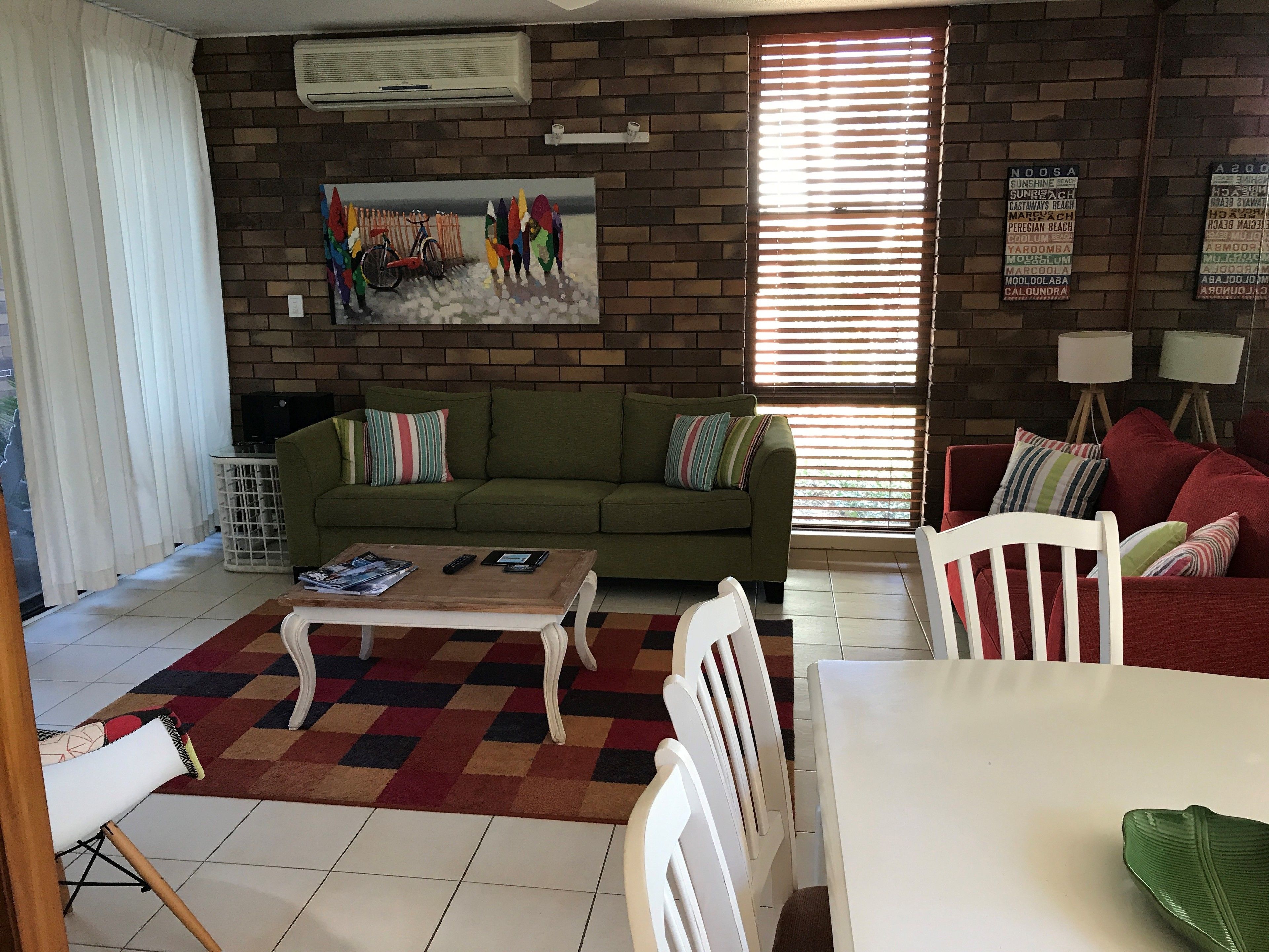 A living room with a green sofa, a coffee table, and a red couch. There is a painting on the wall and a window with blinds.