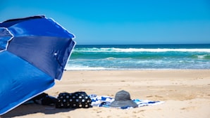 Uma praia com um guarda-sol azul, um chapéu de bolinhas e uma toalha listrada em azul e branco na areia.