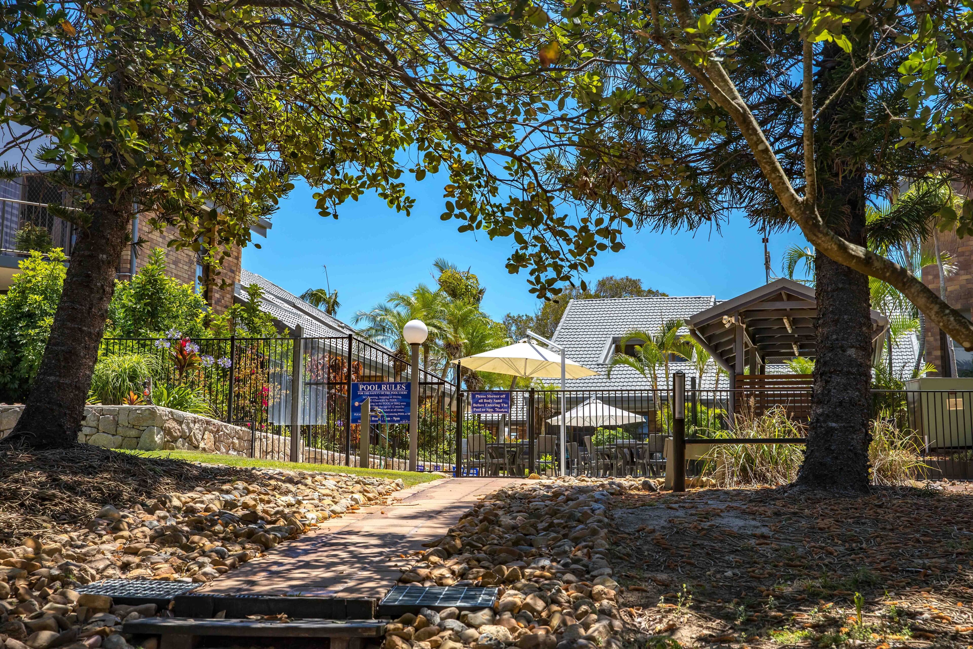 A pathway leading to a pool area with a sign indicating pool rules, surrounded by trees and a stone wall.
