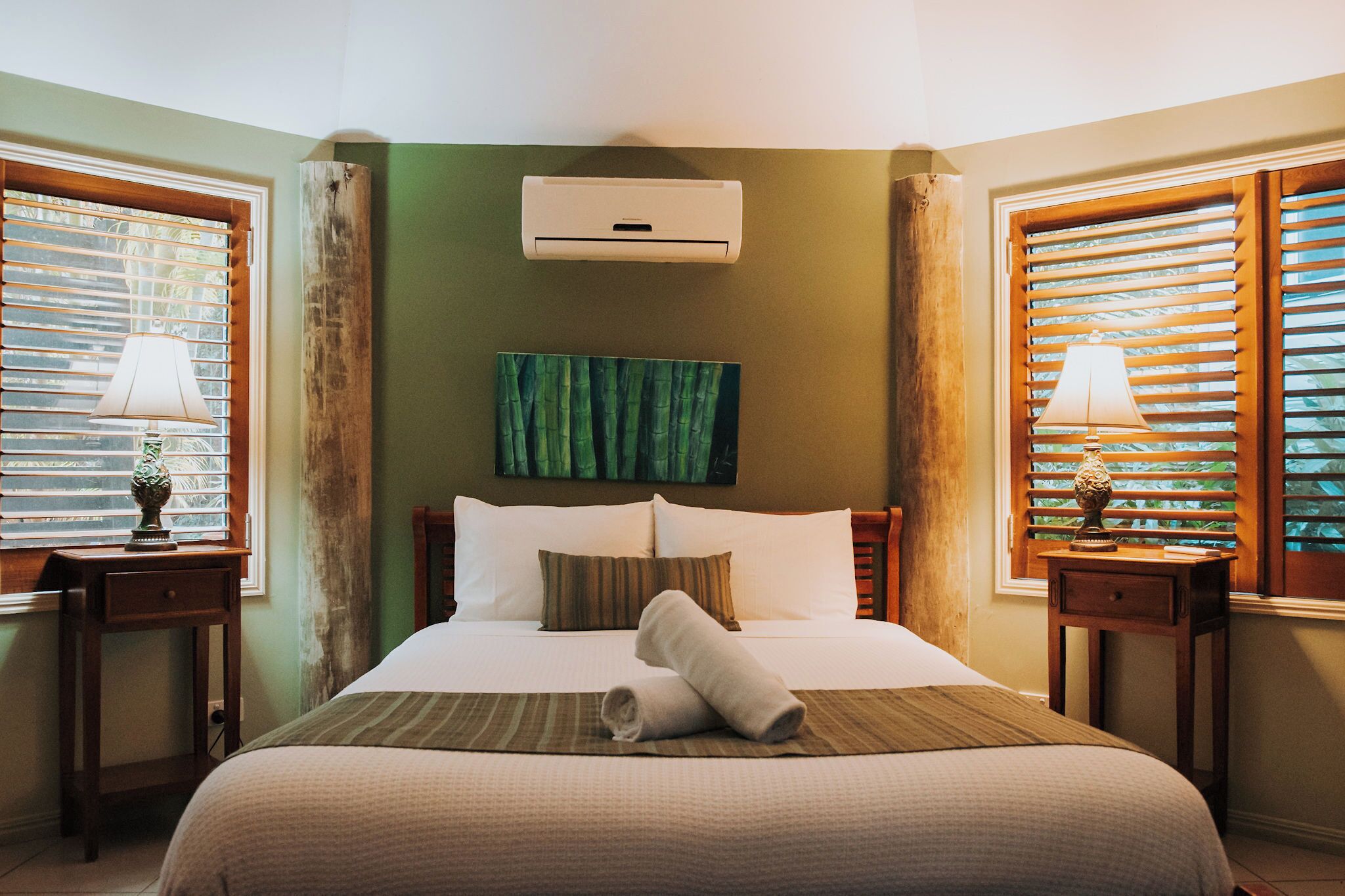 A hotel room with a bed, a wall-mounted air conditioner, a painting, and two nightstands with lamps.