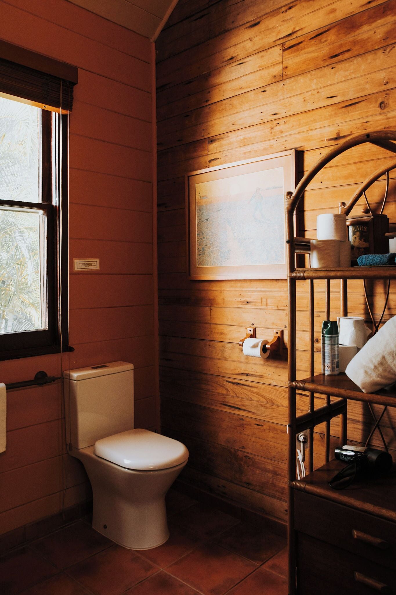 A wooden-paneled bathroom with a toilet, a shelf with toiletries, and a framed picture on the wall.