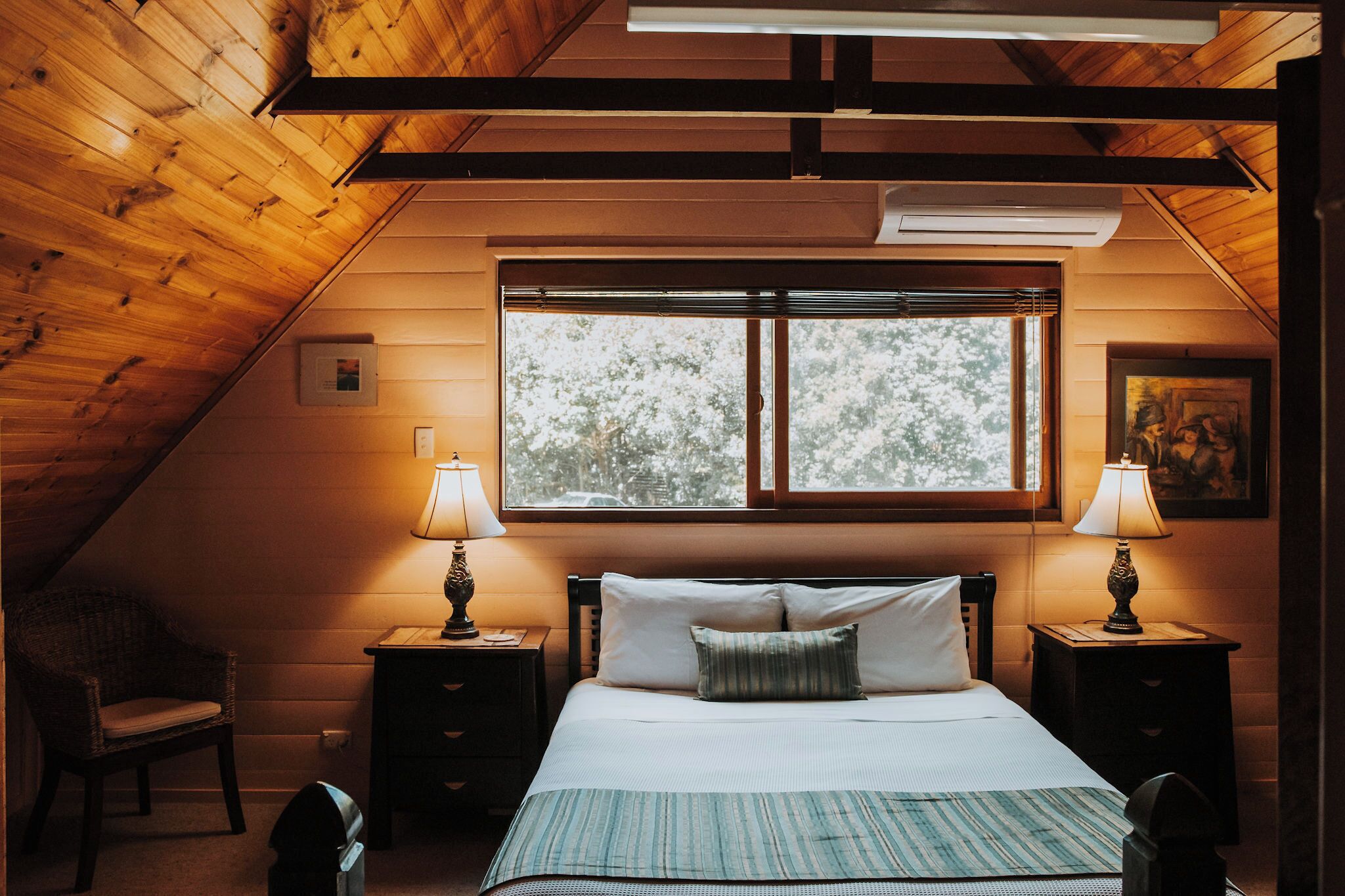 A cozy bedroom with a sloped ceiling, a large window, two bedside tables with lamps, a bed with pillows, and a chair.