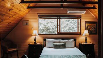 A cozy bedroom with a sloped ceiling, a large window, two bedside tables with lamps, a bed with pillows, and a chair.
