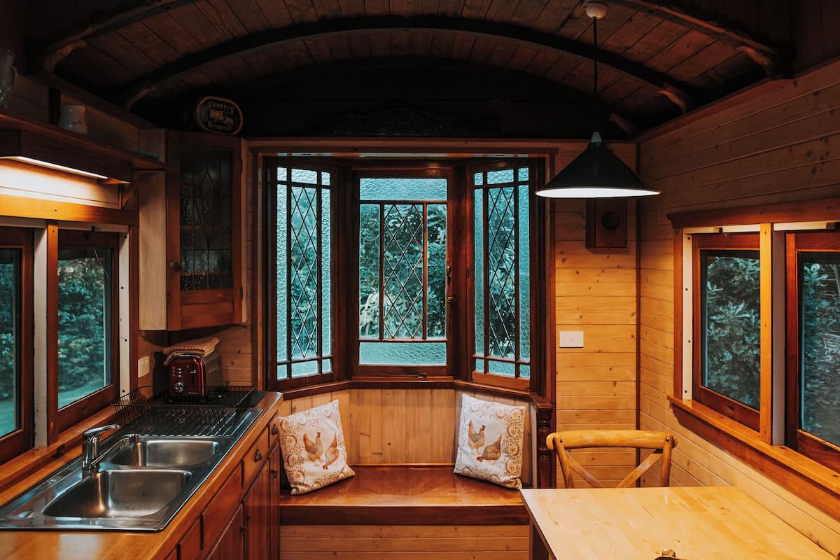 L’intérieur d’un chalet en bois comprend un coin cuisine, un banc avec des coussins et de grandes fenêtres donnant sur la forêt.