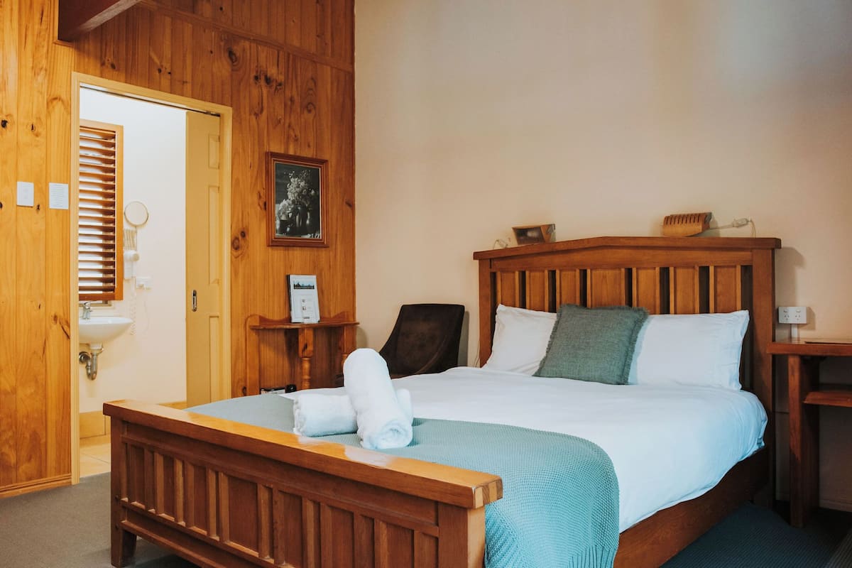 Une pièce aux murs lambrissés, contenant un lit, une table de chevet en bois, une chaise, un banc en bois, une porte en bois et un mur en bois.