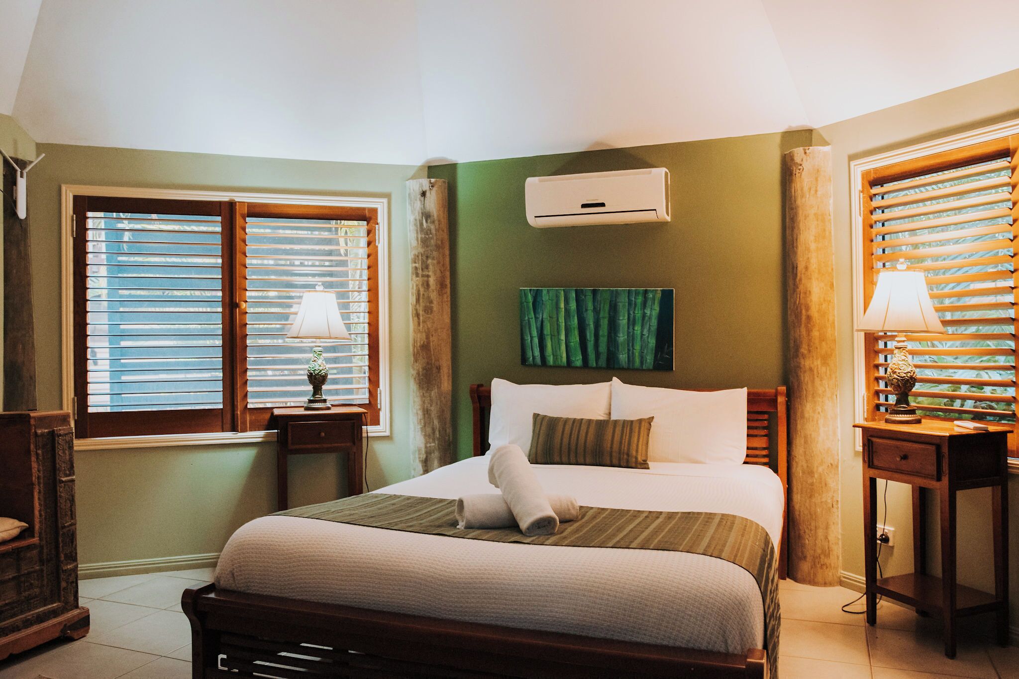 Una habitación de hotel con cama, dos mesitas de noche, aire acondicionado instalado en la pared y una ventana con persianas.
