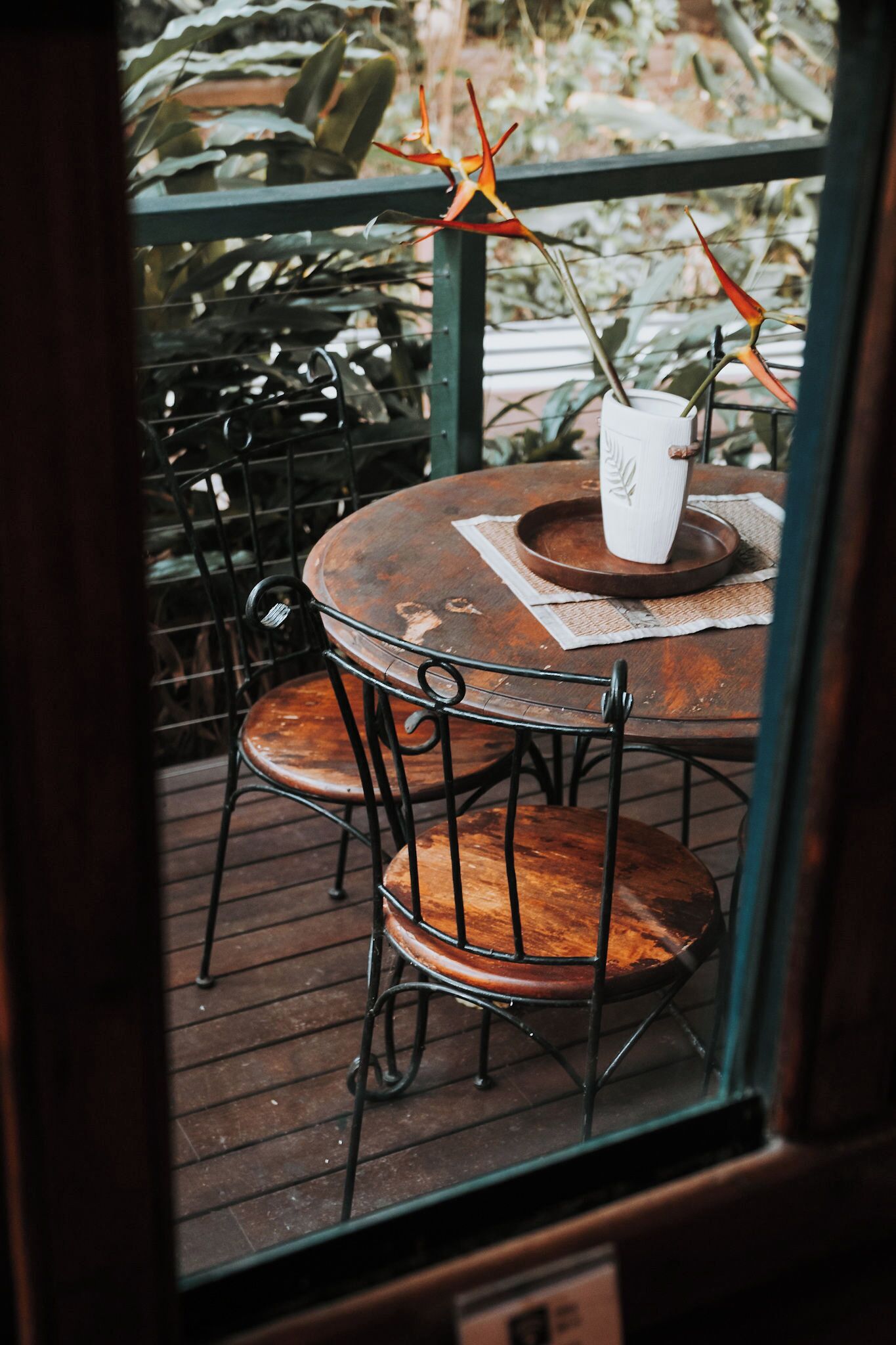 Une table ronde en bois, des chaises en métal, un vase blanc avec une plante et, au fond, une rambarde en métal.