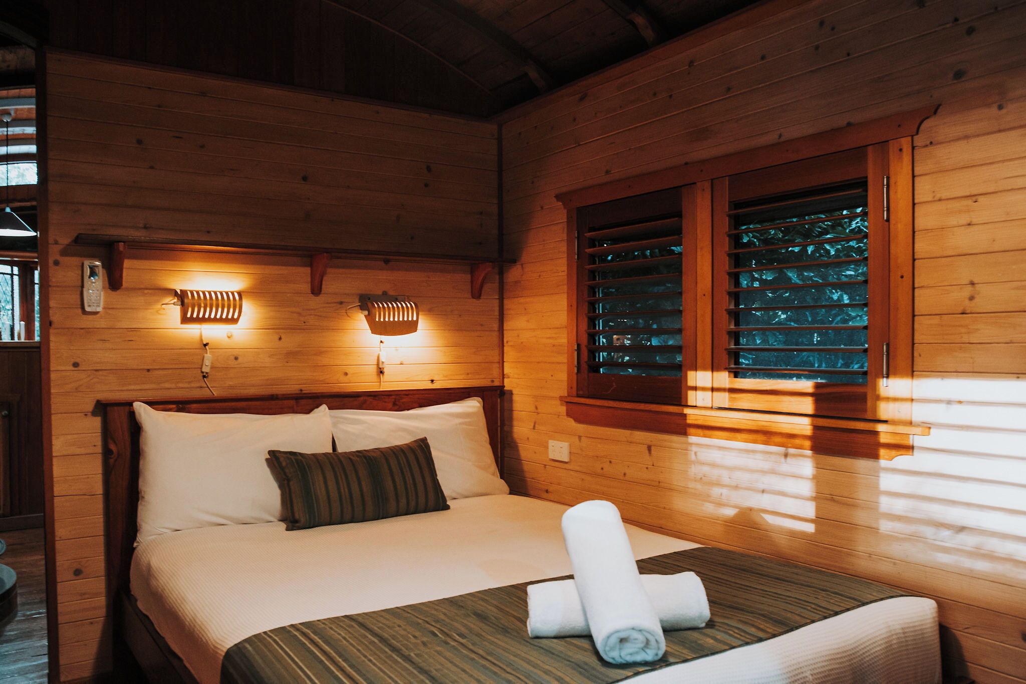 Habitación de cabaña de madera con cama, dos luces fijadas a la pared, una ventana con persianas y una pequeña mesa con una toalla enrollada.