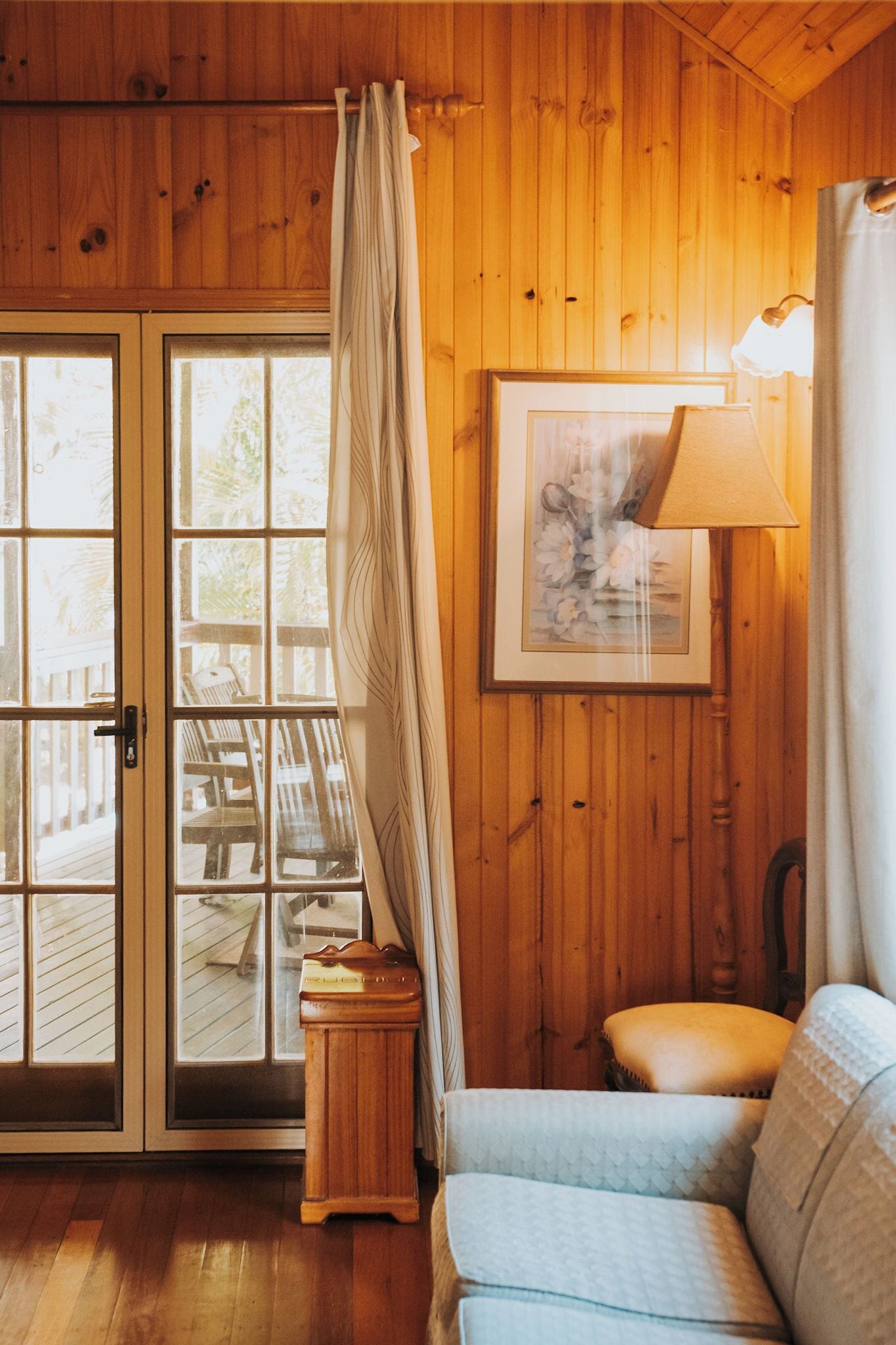 A wooden interior with a sofa, a framed picture, a lamp, and a door leading to an outdoor area.