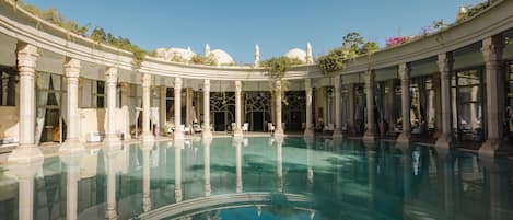 A large indoor swimming pool with classical architecture, including columns and arches, surrounded by seating areas.