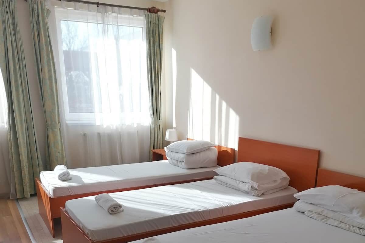 A hotel room with two single beds, a window with curtains, a nightstand, and a wall-mounted lamp.