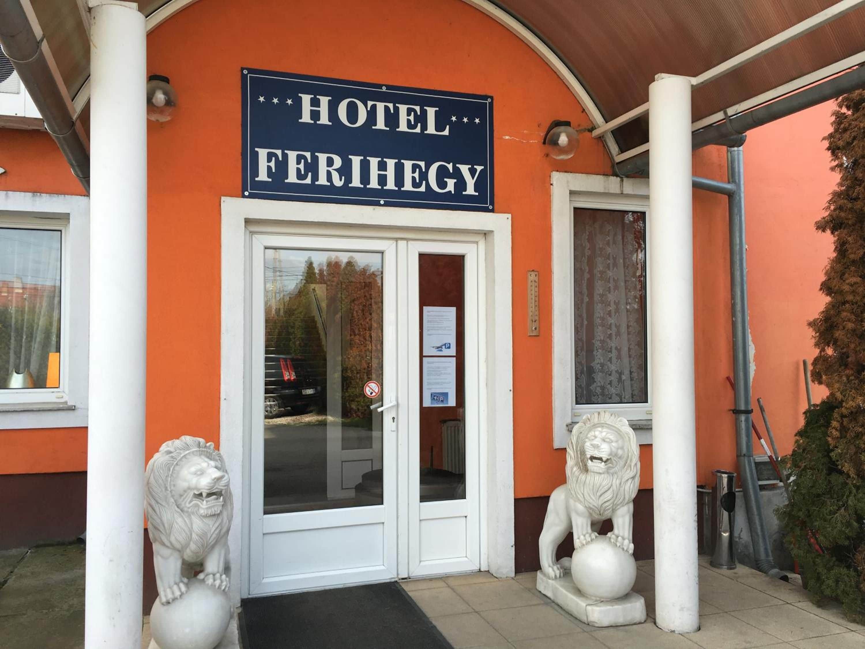 A hotel entrance with a sign reading 'HOTEL FERIHEGY,' two lion statues, and a covered entrance area.