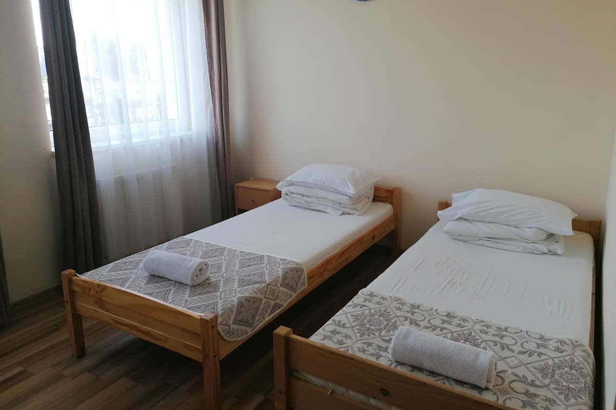 Two single beds with wooden frames and patterned bedspreads, a small wooden nightstand, and a window with sheer curtains.