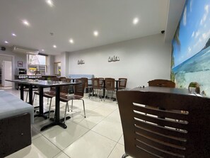 A modern dining area with a long table and multiple chairs, a kitchen area with a 'Good Morning' sign, and a mural of a beach scene on the wall. - Trebovir Hotel (London)