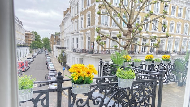 Ein Balkon mit schmiedeeisernen Geländern, Blumentöpfen und Blick auf eine Straße mit geparkten Autos und Gebäuden.