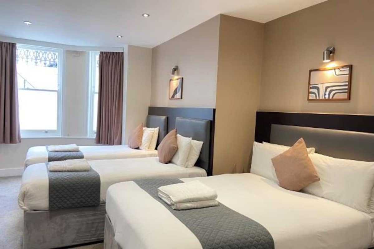 Une chambre d’hôtel avec trois lits, chacun doté d’une tête de lit, d’une grande fenêtre avec des rideaux et d’un éclairage fixé au mur.