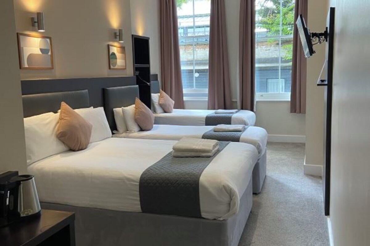 A hotel room with two beds, a bedside table, a wall-mounted TV, and a window with curtains.