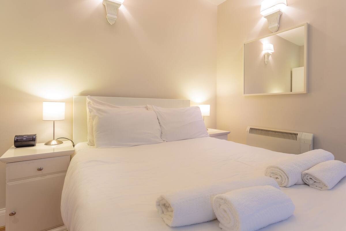 A neatly made bed with white linens, two bedside lamps, a bedside table with a lamp, a mirror, and a radiator.