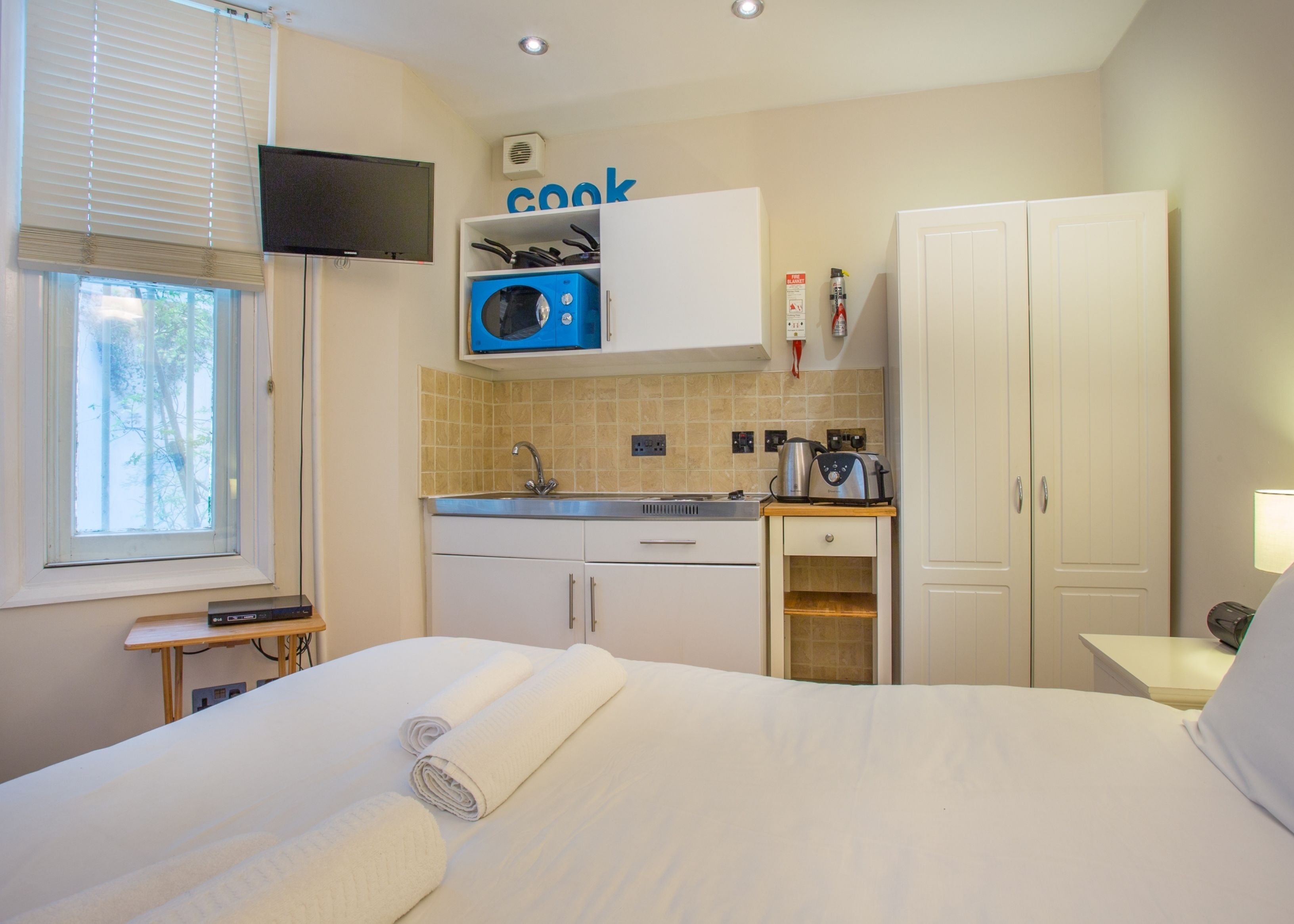 A compact hotel room with a bed, a small kitchenette, a TV, and a window with blinds.