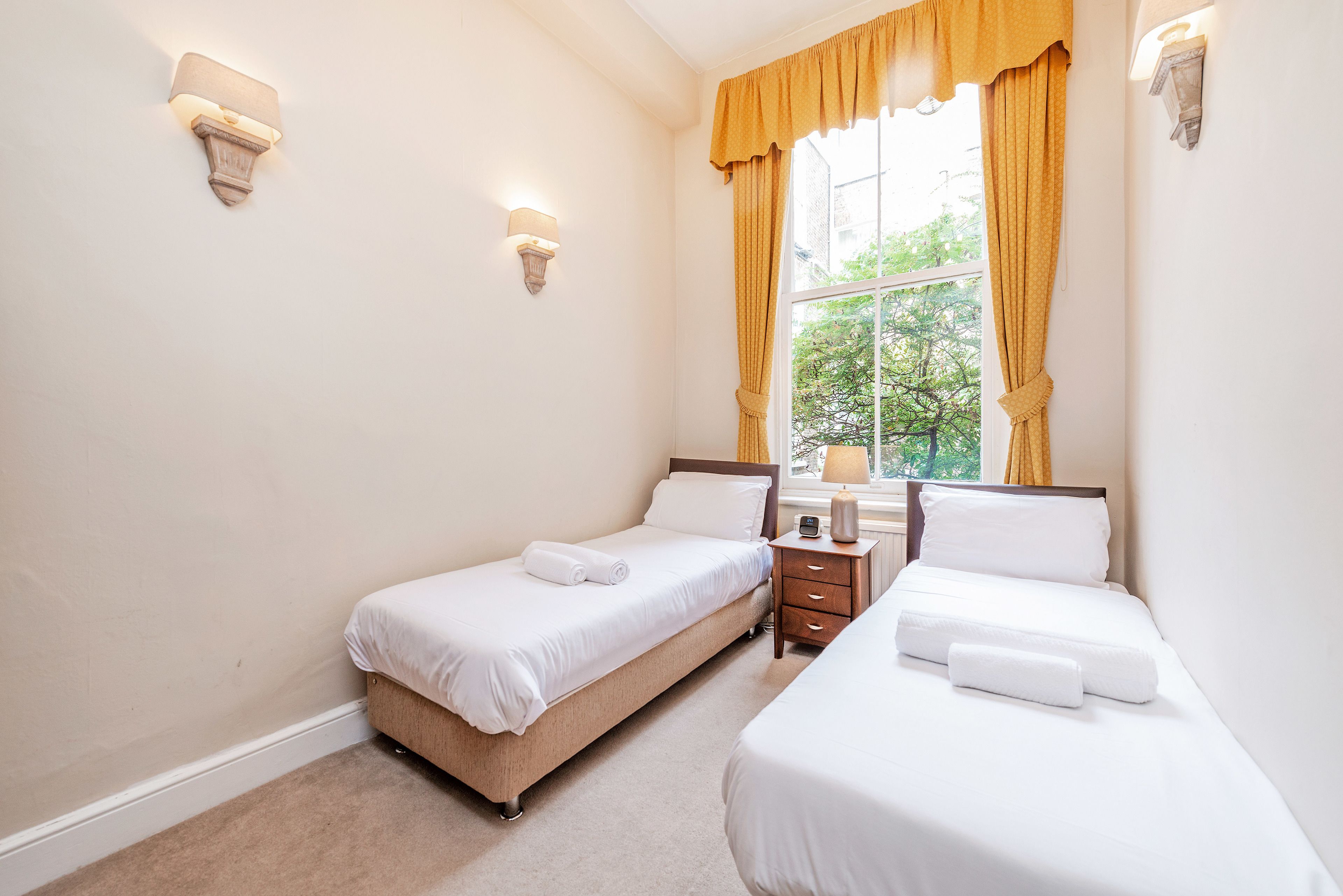 A hotel room with two beds, a nightstand, a lamp, and a window with curtains.