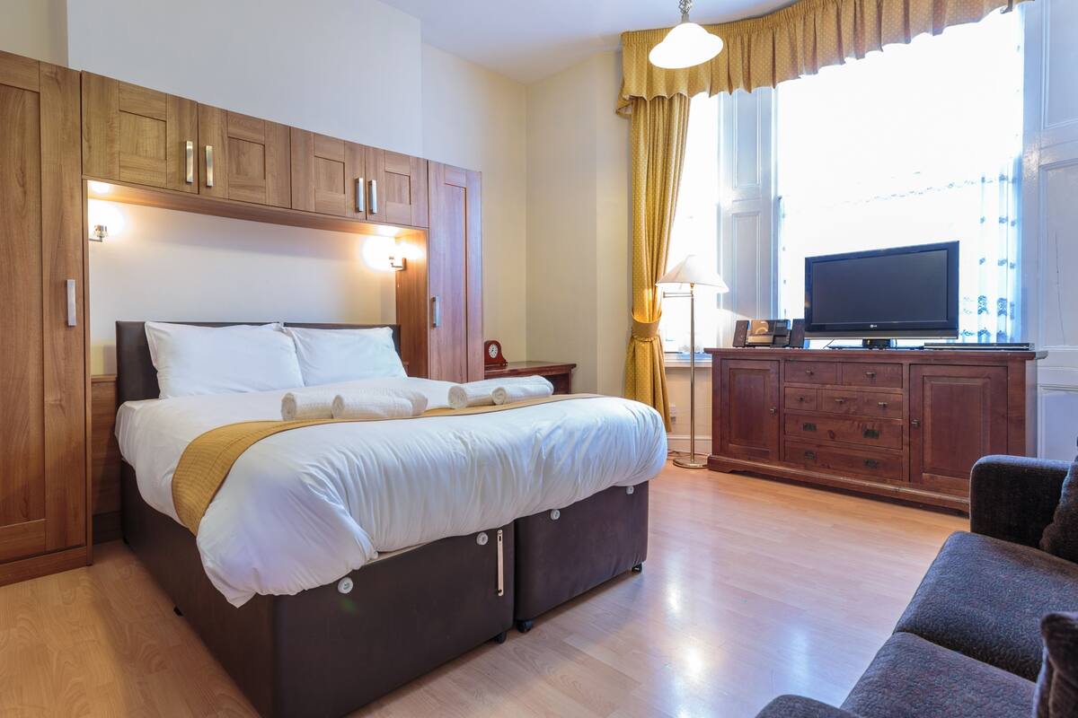 A modern hotel room with a large bed, wooden wardrobe, a flat-screen TV, and a sofa.