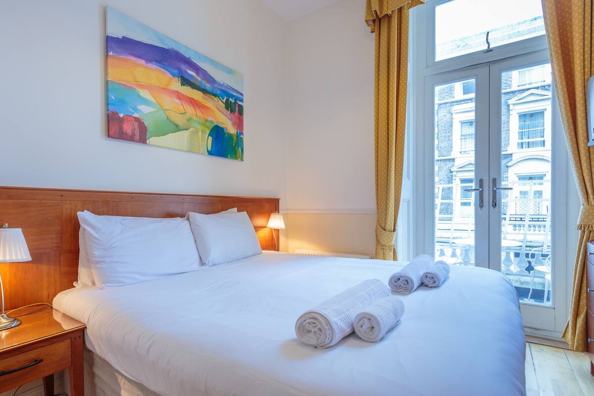 A hotel room with a large bed, a wooden headboard, a painting on the wall, and a window with curtains.