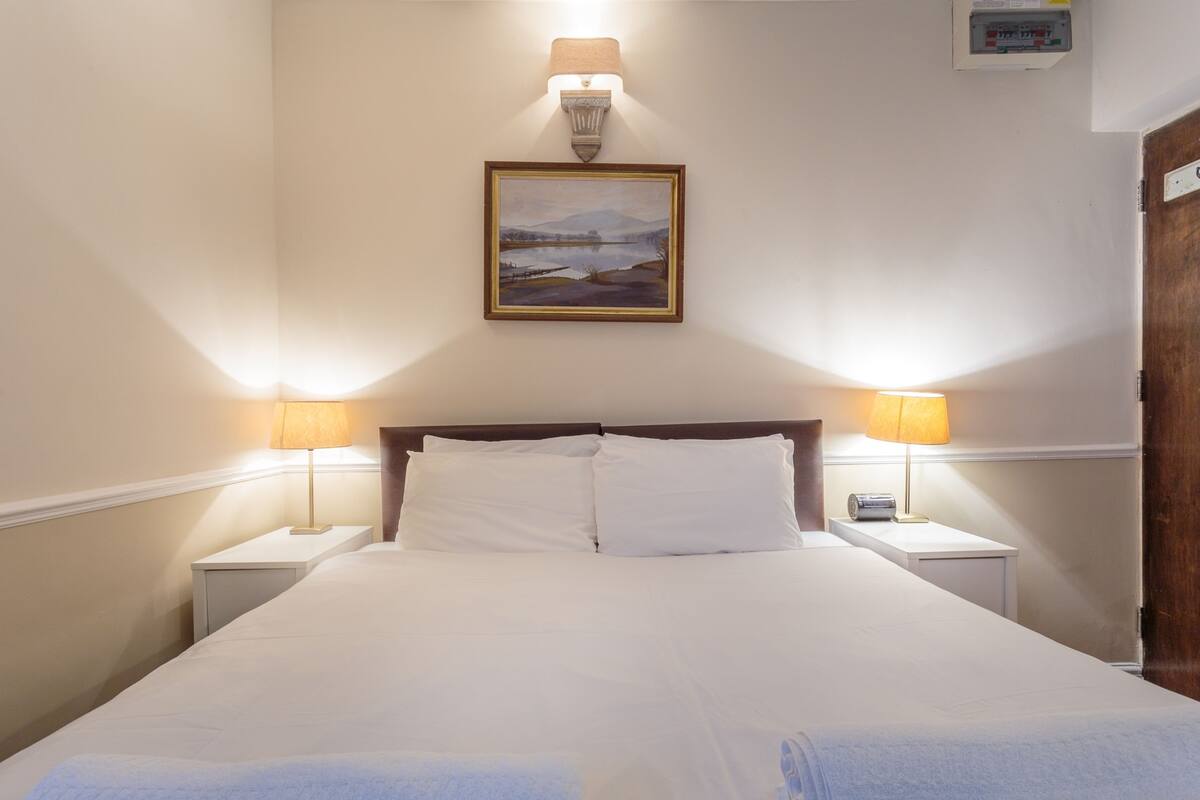 A neatly made bed with white linens, two bedside lamps, a framed landscape painting, and a wooden door.