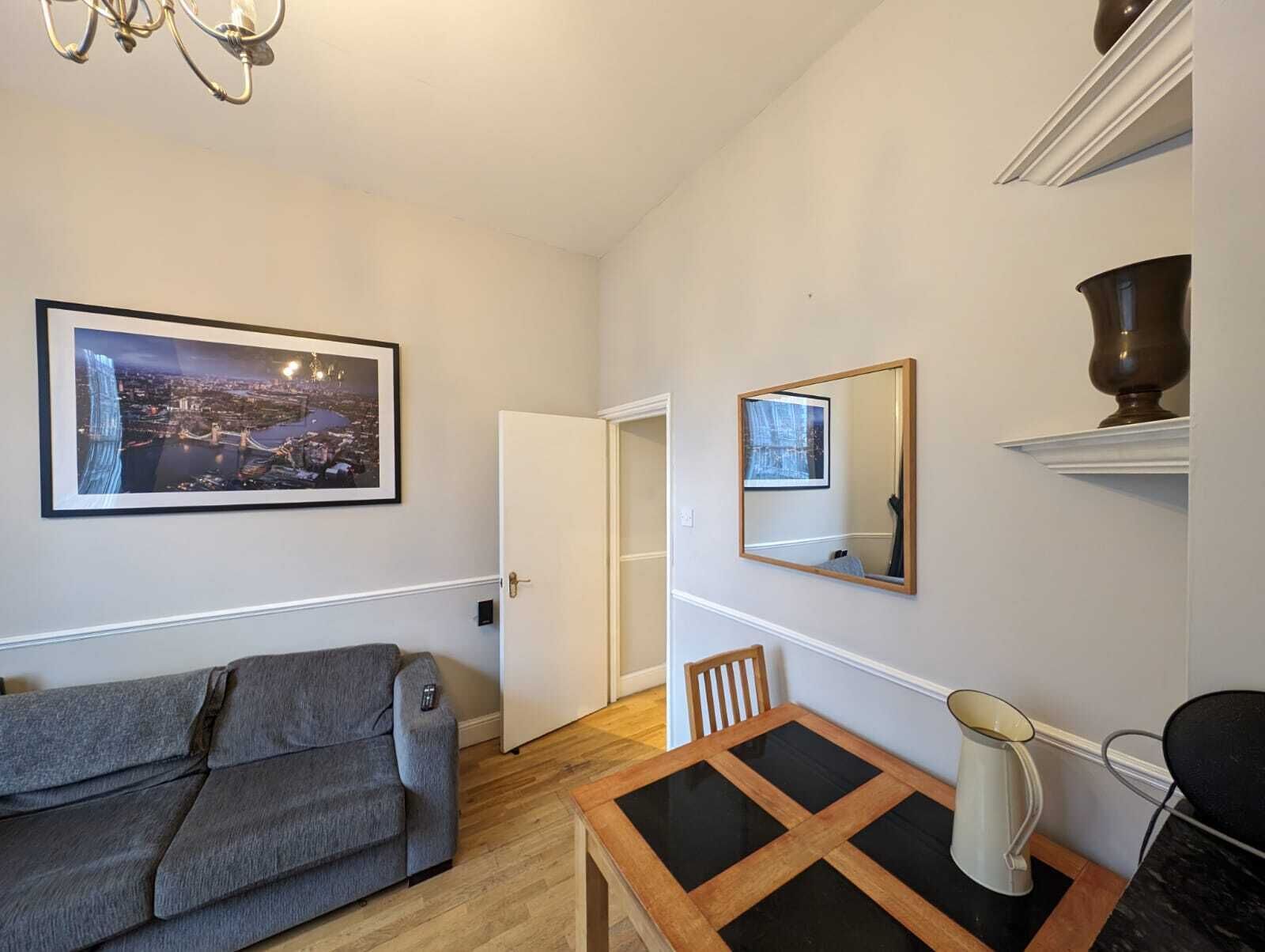 Ett modernt vardagsrum med en grå soffa, ett matbord med glasskiva, en spegel och ett inramat fotografi av en stadsbild på väggen.