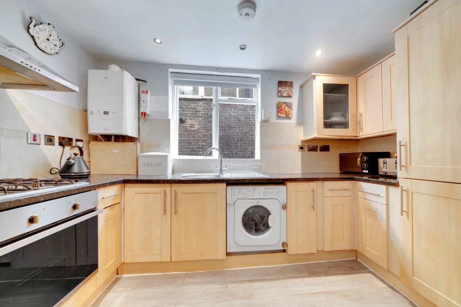 A modern kitchen with wooden cabinets, a built-in oven, a washing machine, and a window with a view of the city.