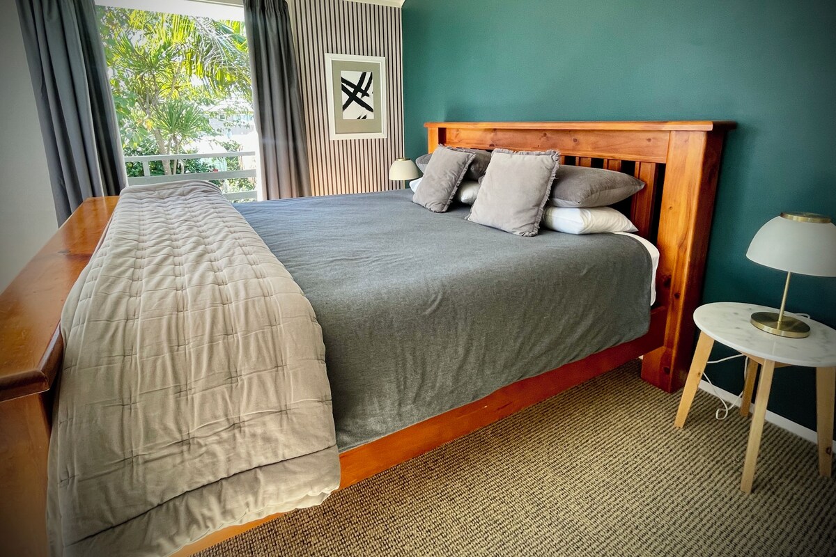 Una cama bien tendida con colcha y almohadas grises, cabecera de madera, mesita de noche con lámpara y vista a zonas verdes por la ventana.
