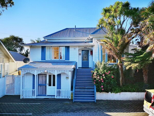 Una casa de dos pisos con techo azul y paredes blancas, un porche con barandilla blanca y una escalera que lleva a la puerta principal.