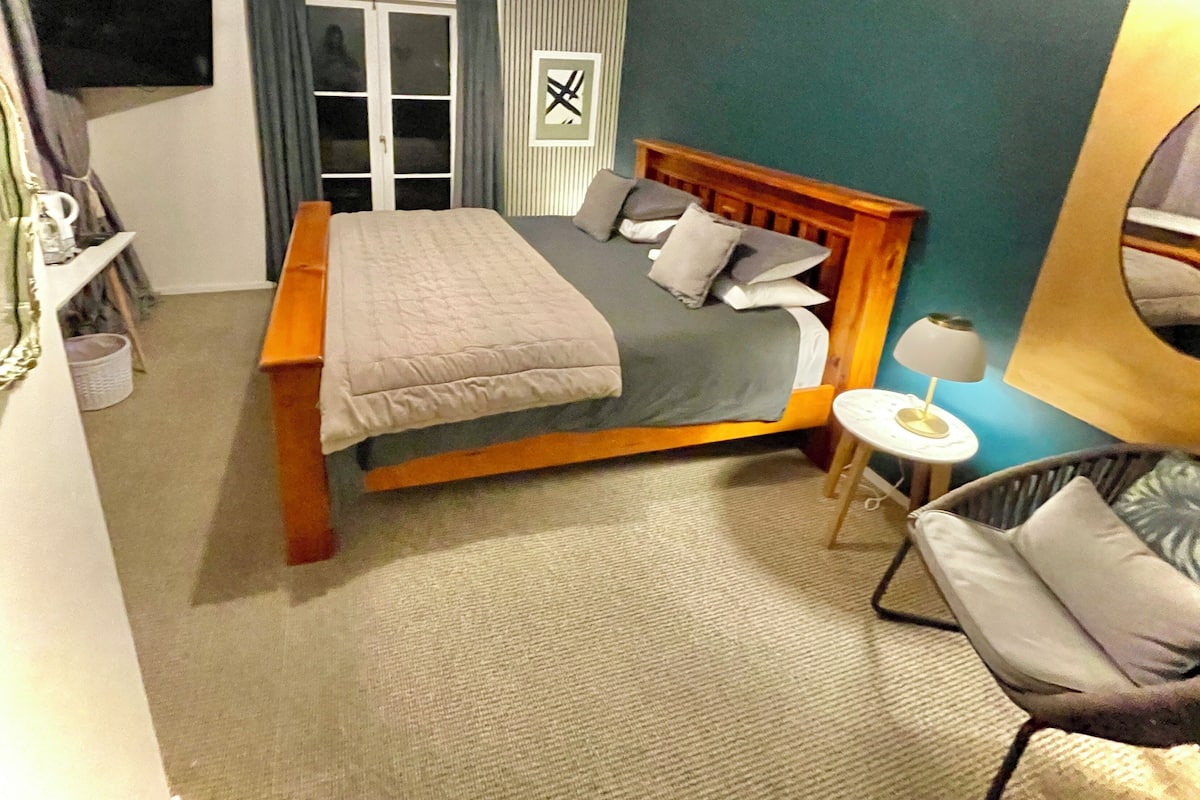 Un dormitorio con una cama, una silla, una mesita, un televisor y una ventana con cortinas.