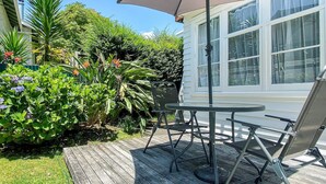 A patio with a table and chairs, surrounded by lush greenery and a white house. - Bavaria Boutique Hotel (Auckland)