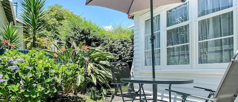 Une terrasse avec une table et des chaises, entourée d’une végétation luxuriante et d’une maison blanche.