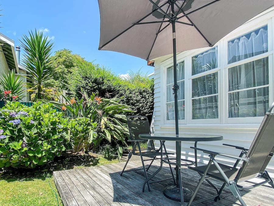 A patio with a table and chairs, surrounded by lush greenery and a white house.
