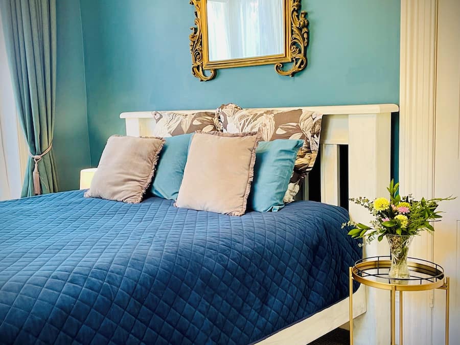 A bedroom with a bed, a round gold side table, a mirror, and a vase with flowers.