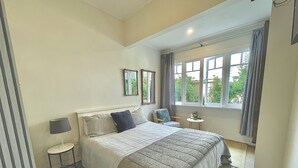 A bedroom with a large bed, a small table, a chair, and a window with curtains. - Bavaria Boutique Hotel (Auckland)