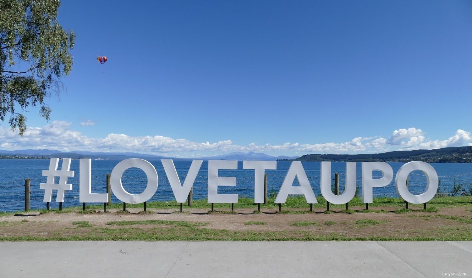 A large sign with the text "#LOVETAUPO" in white letters against a backdrop of a clear blue sky and a body of water.