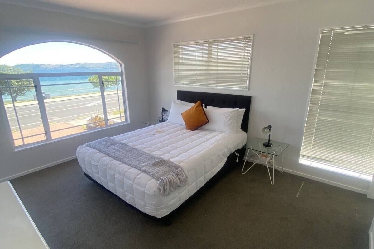 A bedroom with a large bed, a small bedside table, a window with a view of a body of water, and a ceiling light.