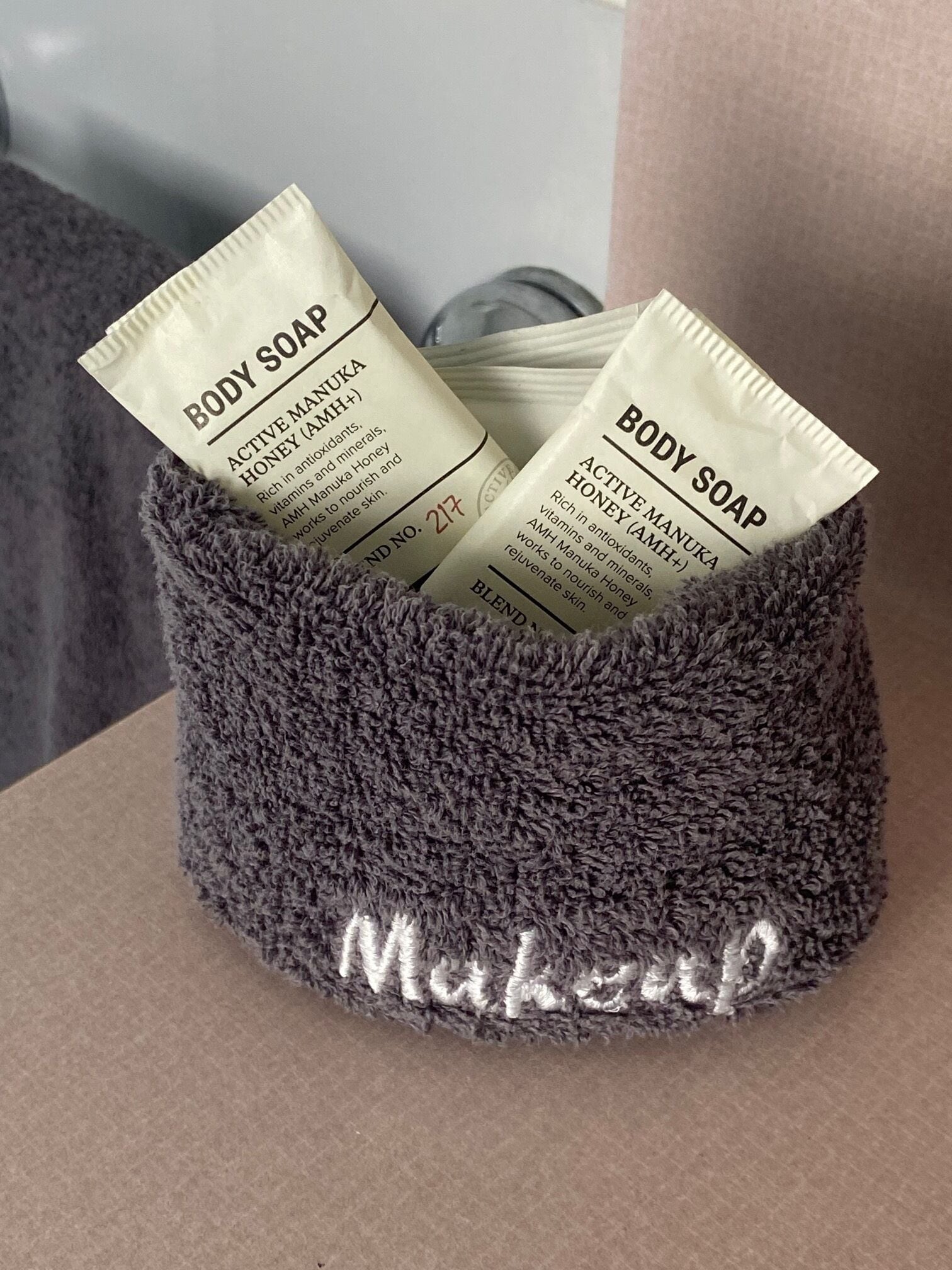 A grey, textured container with the word "Makeup" on it, holding two packets of body soap labeled "Active Manuka Honey (AMH+)."