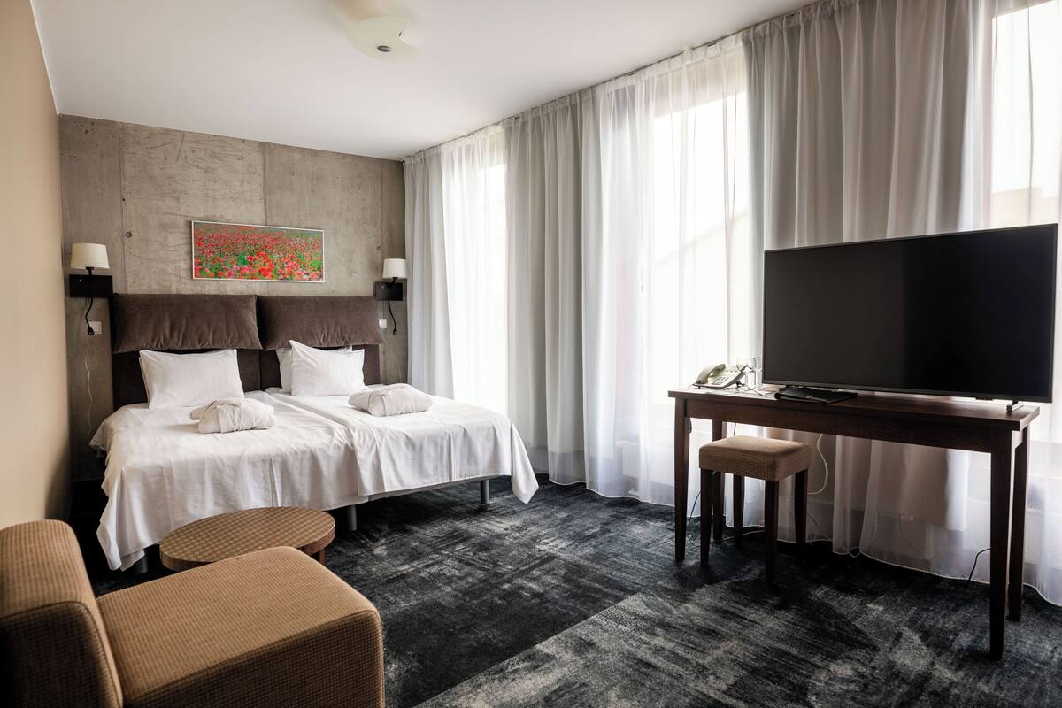 A hotel room with a bed, a desk with a TV, a chair, and a large window with curtains.