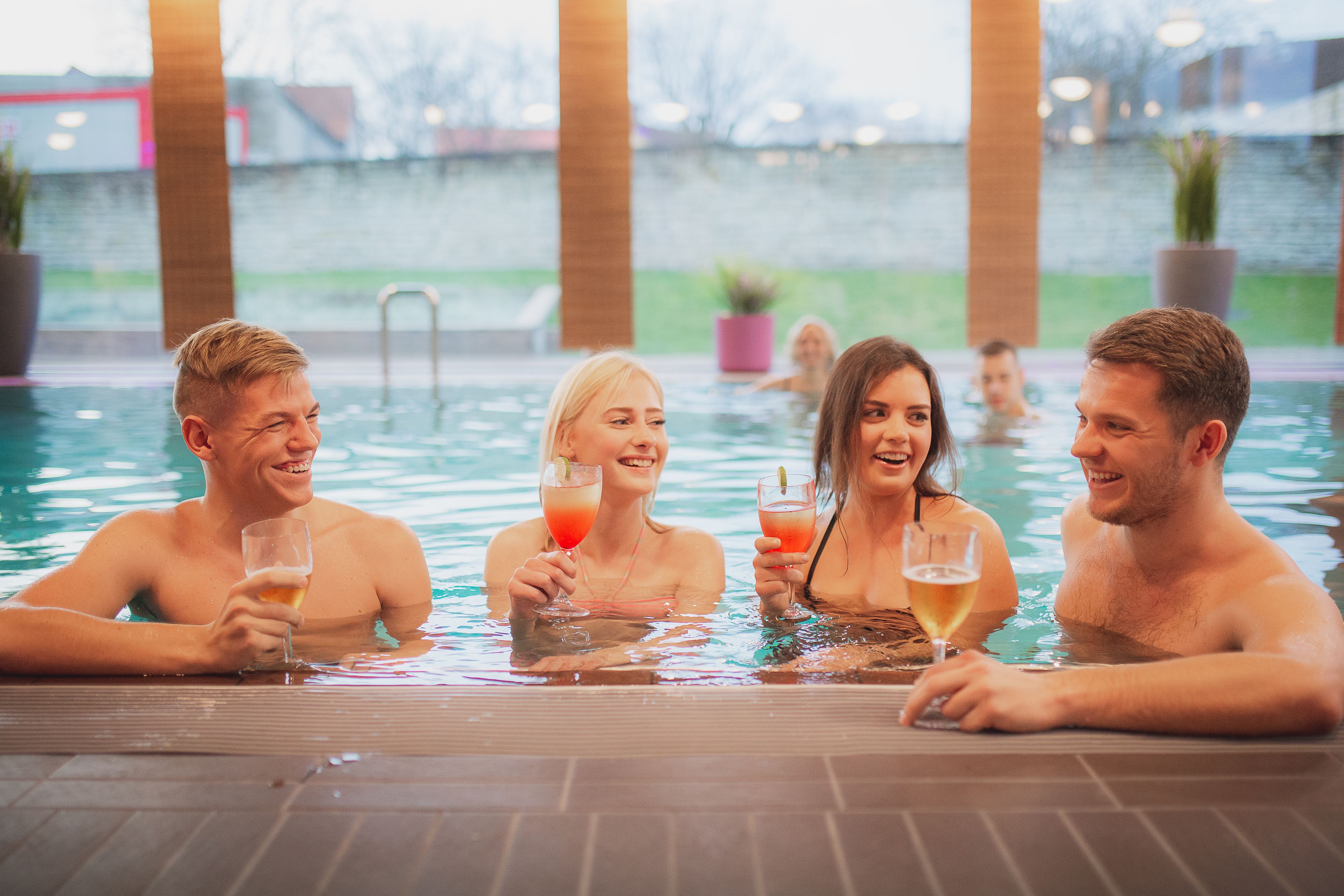 Un grupo de personas en una piscina cubierta con agua caliente, disfrutando de bebidas, rodeadas de plantas en macetas y un muro de ladrillo al fondo.