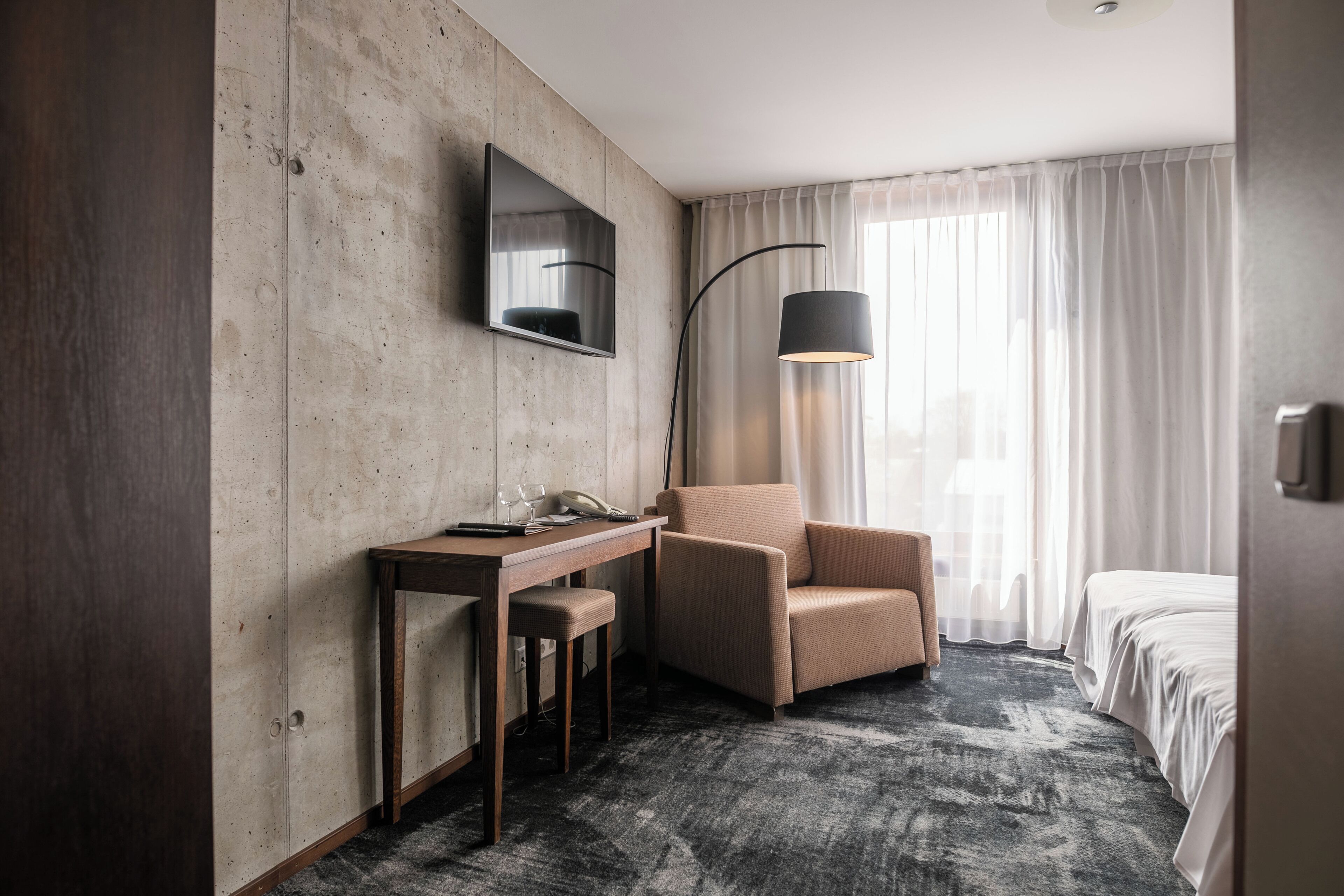 Ein Hotelzimmer mit einem Holztisch, einem Stuhl, einem Bett, einem an der Wand befestigten Fernseher und einer Stehlampe.