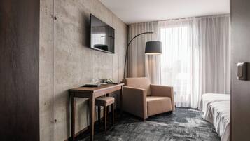 A hotel room with a wooden desk, a chair, a bed, a TV mounted on the wall, and a floor lamp.