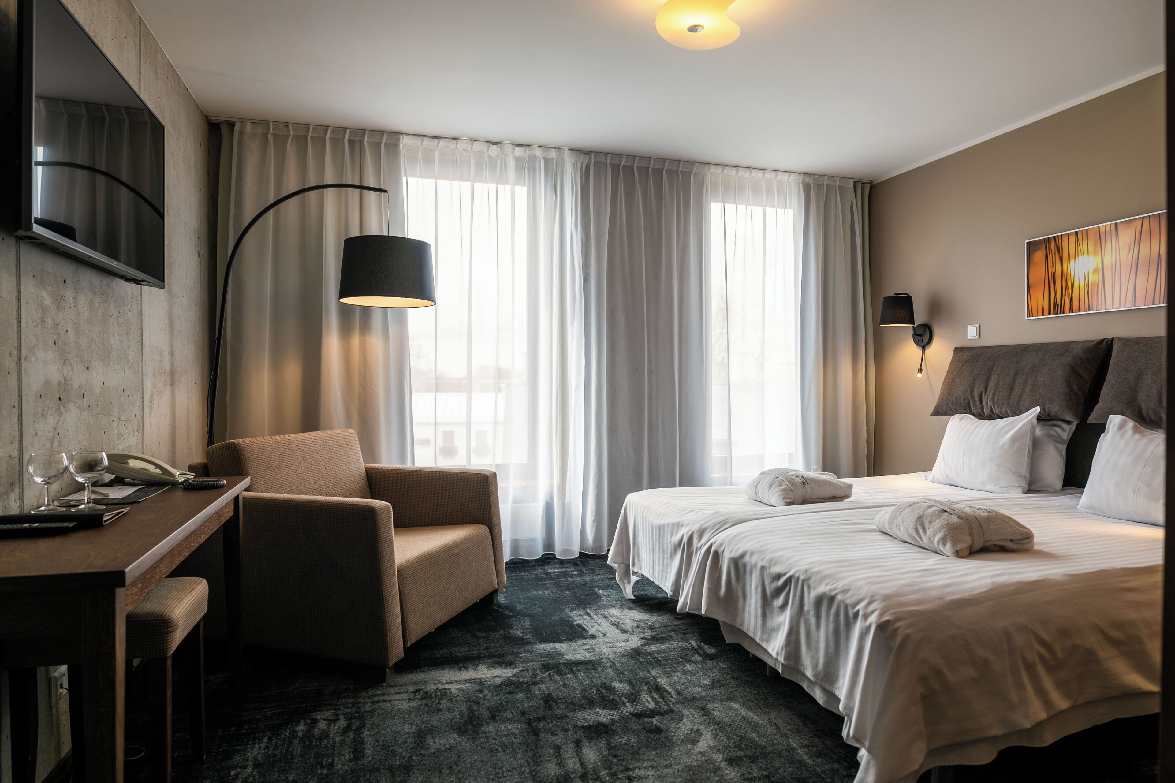 Habitación de hotel con cama, escritorio, silla, televisor y una ventana grande con cortinas.