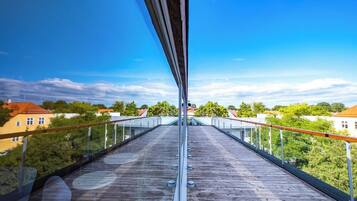 A wooden bridge with glass railings, leading to a view of buildings and trees.