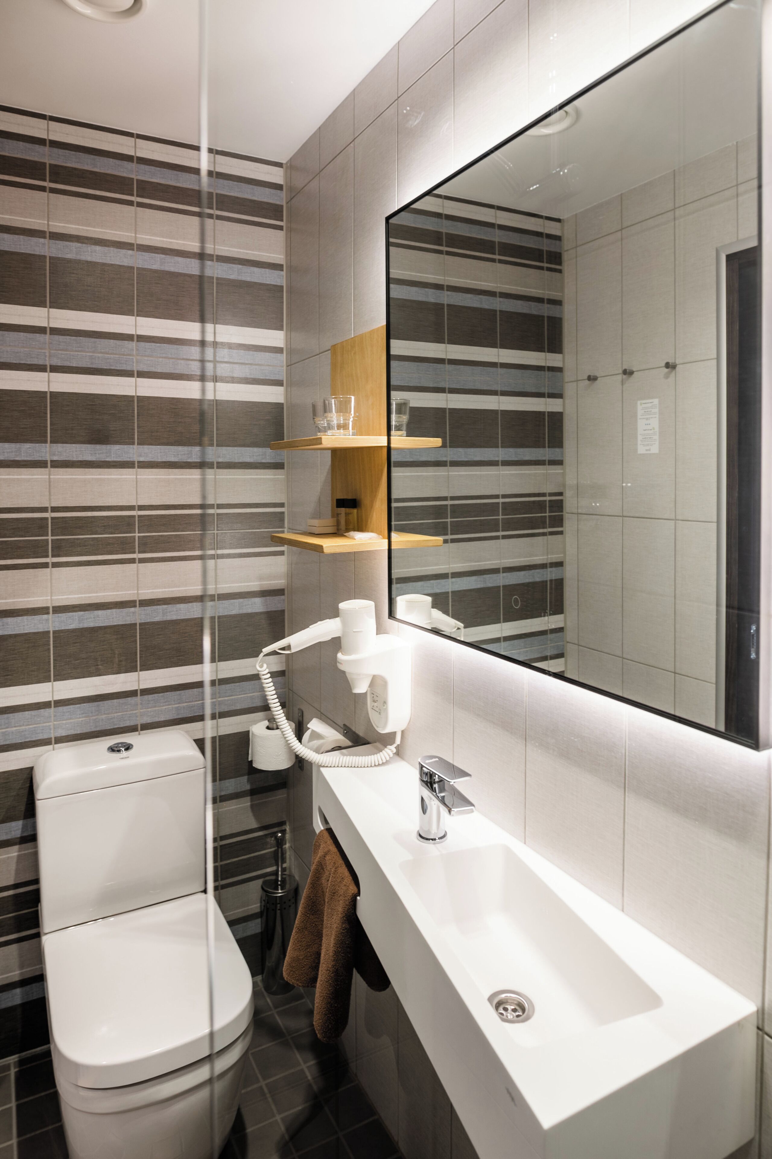 Un baño moderno con inodoro, lavamanos y espejo. Las paredes tienen azulejos a rayas y hay un estante de madera sobre el lavamanos.