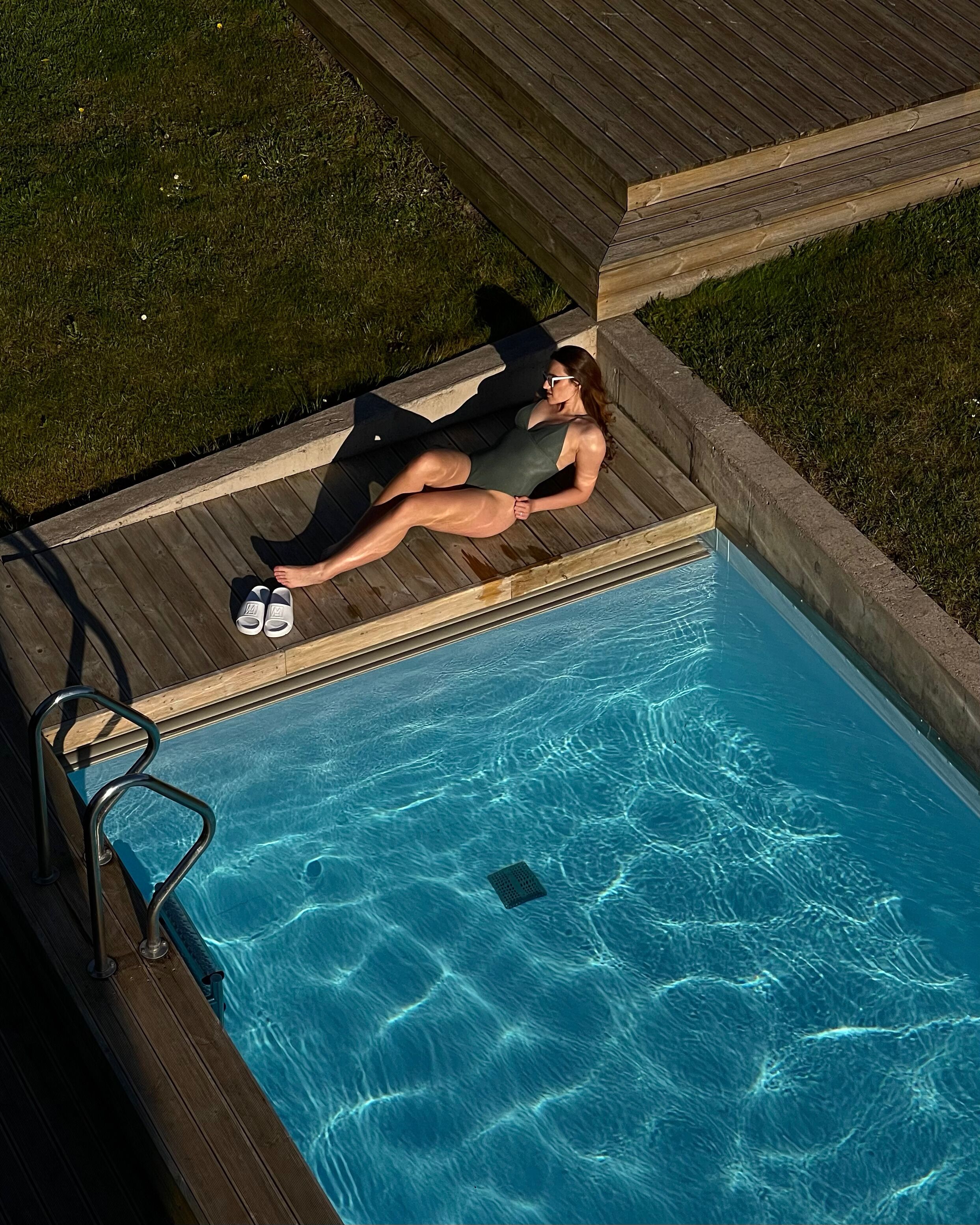 A person relaxing on a wooden deck by a swimming pool.