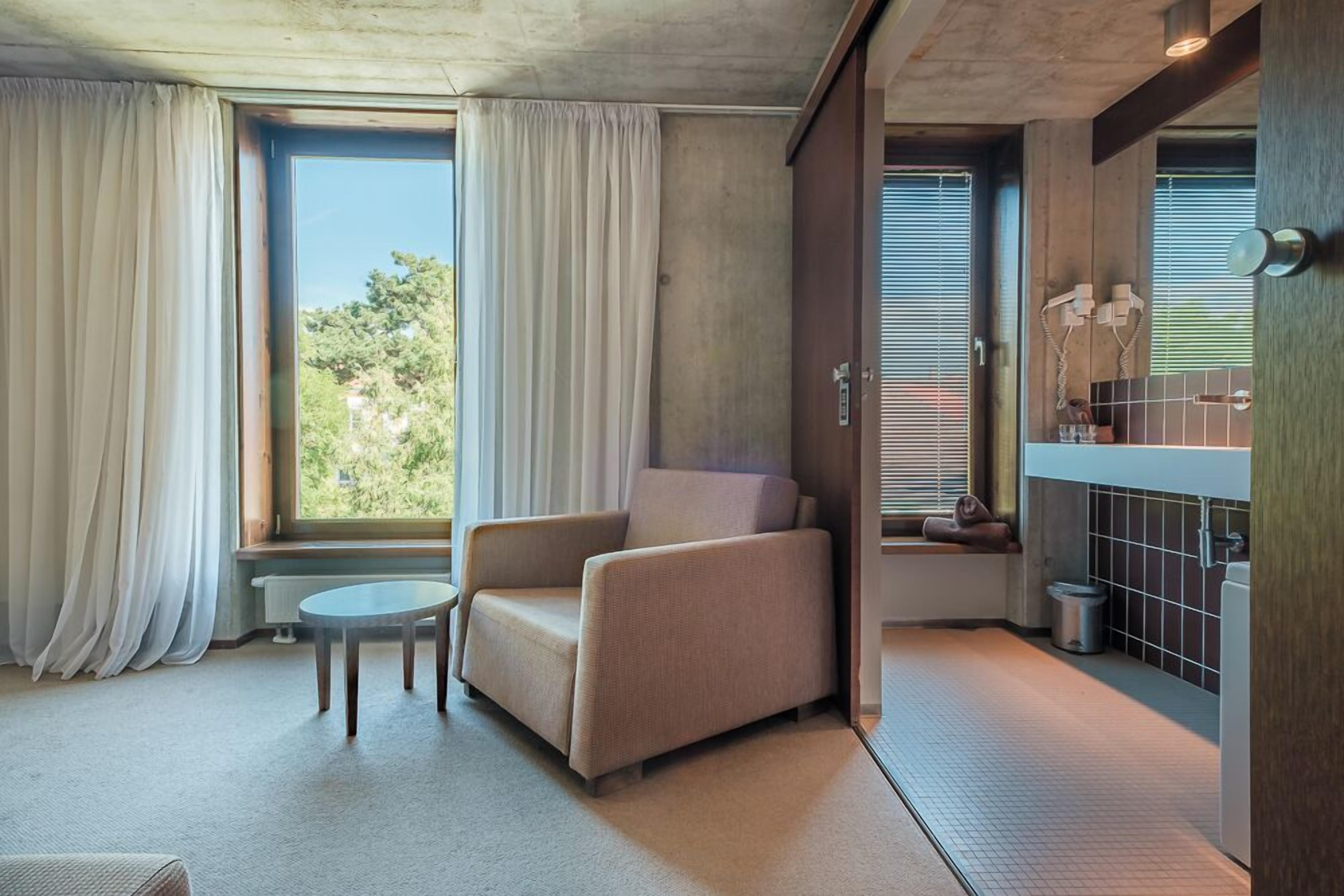 Quarto moderno com poltrona bege, mesa redonda pequena, janela com cortinas e área de cozinha compacta com pia e fogão.